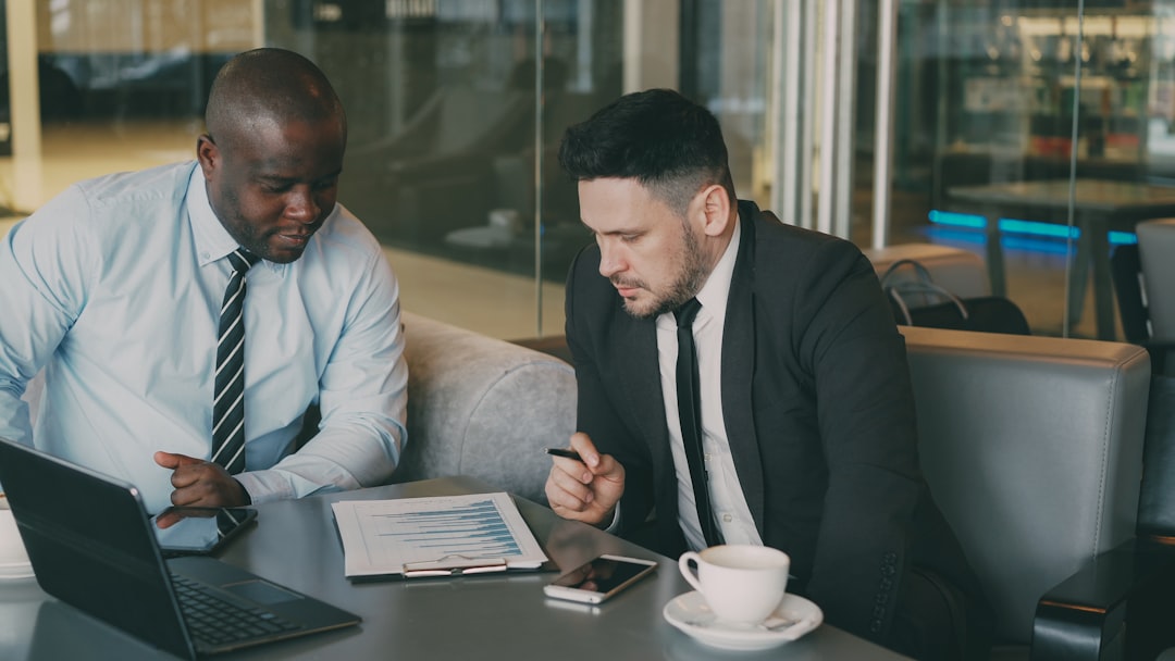Two businessmen in formal clothes sitting and discussing their business financial charts while looking at notepad in glassy modern cafe during lunch time